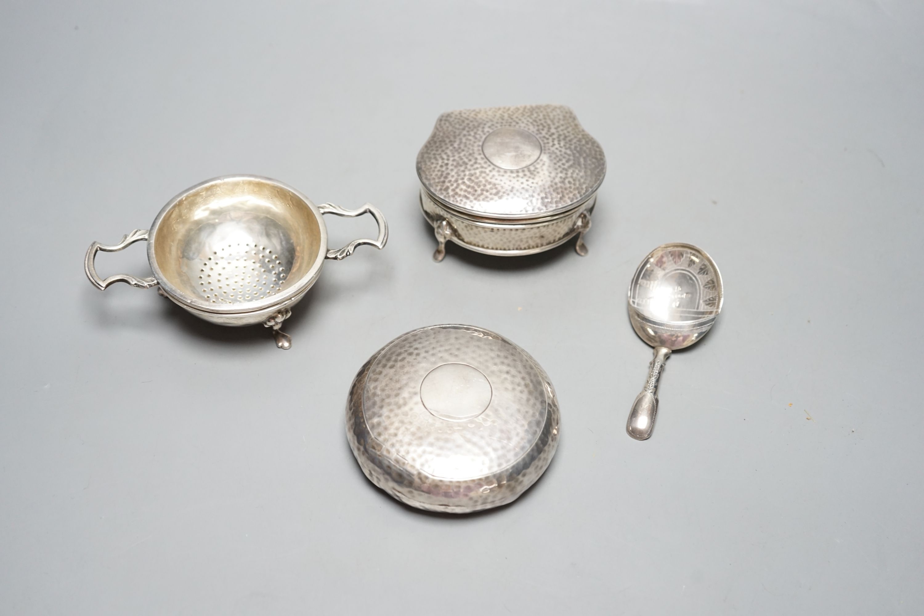 An Edwardian planished silver circular tobacco box, by George Unite, Birmingham, 1902, 79mm, a similar silver trinket box, a George III silver caddy spoons, Birmingham, 1814 and a modern silver teas strainer on stand.